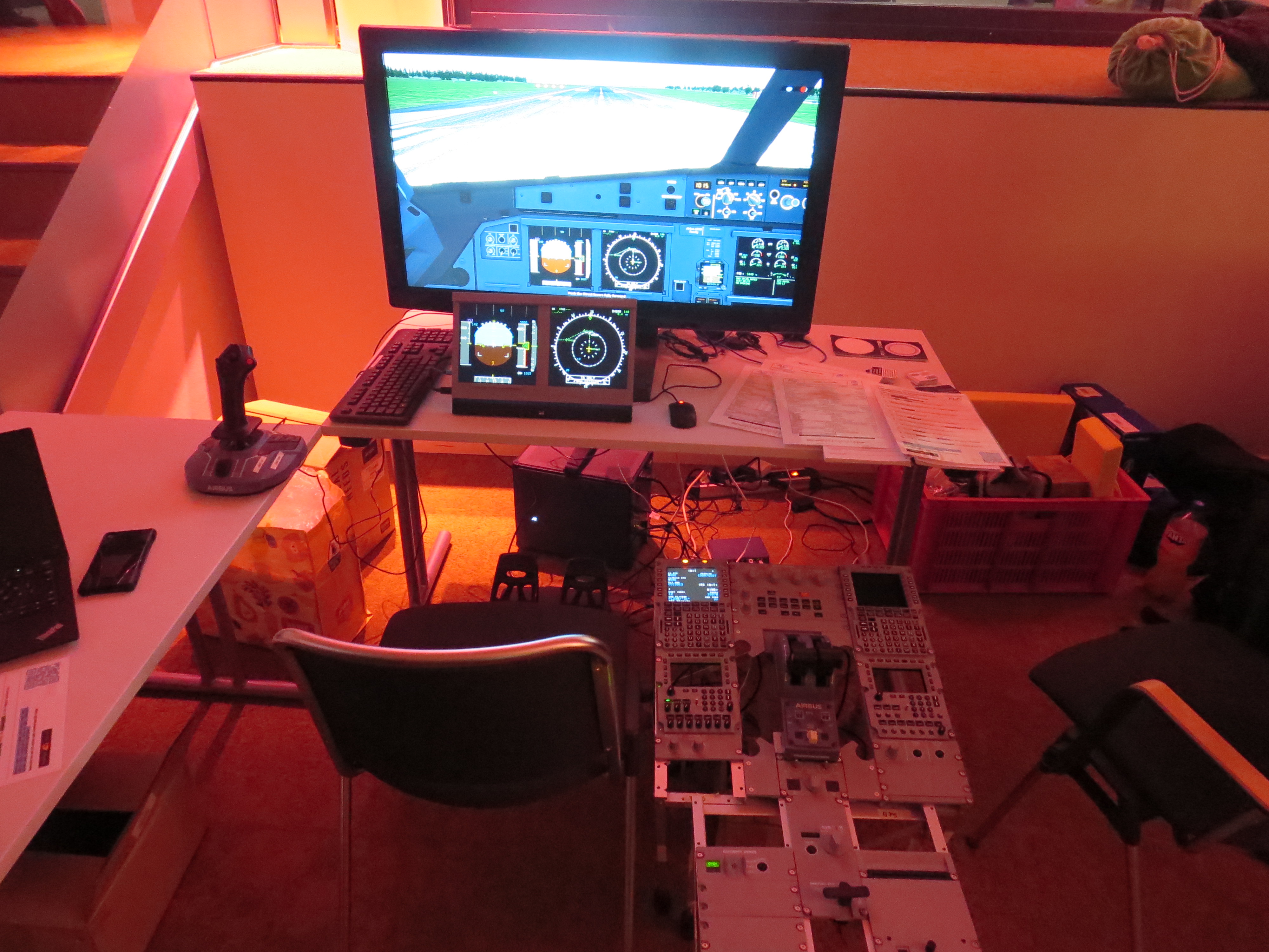 The setup consists of two tables arranged in an L shape, one behind it and one to the site where the side stick rests on. On the table in the back is a big monitor showing the simulator and a smaller one, showing the primary flight display and navigation display. In front of the monitors is a chair for the pilot flying, to the right of it is the DIY pedestal made out of many 3d printed panels with lots of buttons, knobs and switches. To the right of that is a second chair for a pilot monitoring. Behind the desk there are some boxes which were used to transport the sim as well as the PC running the simulation.